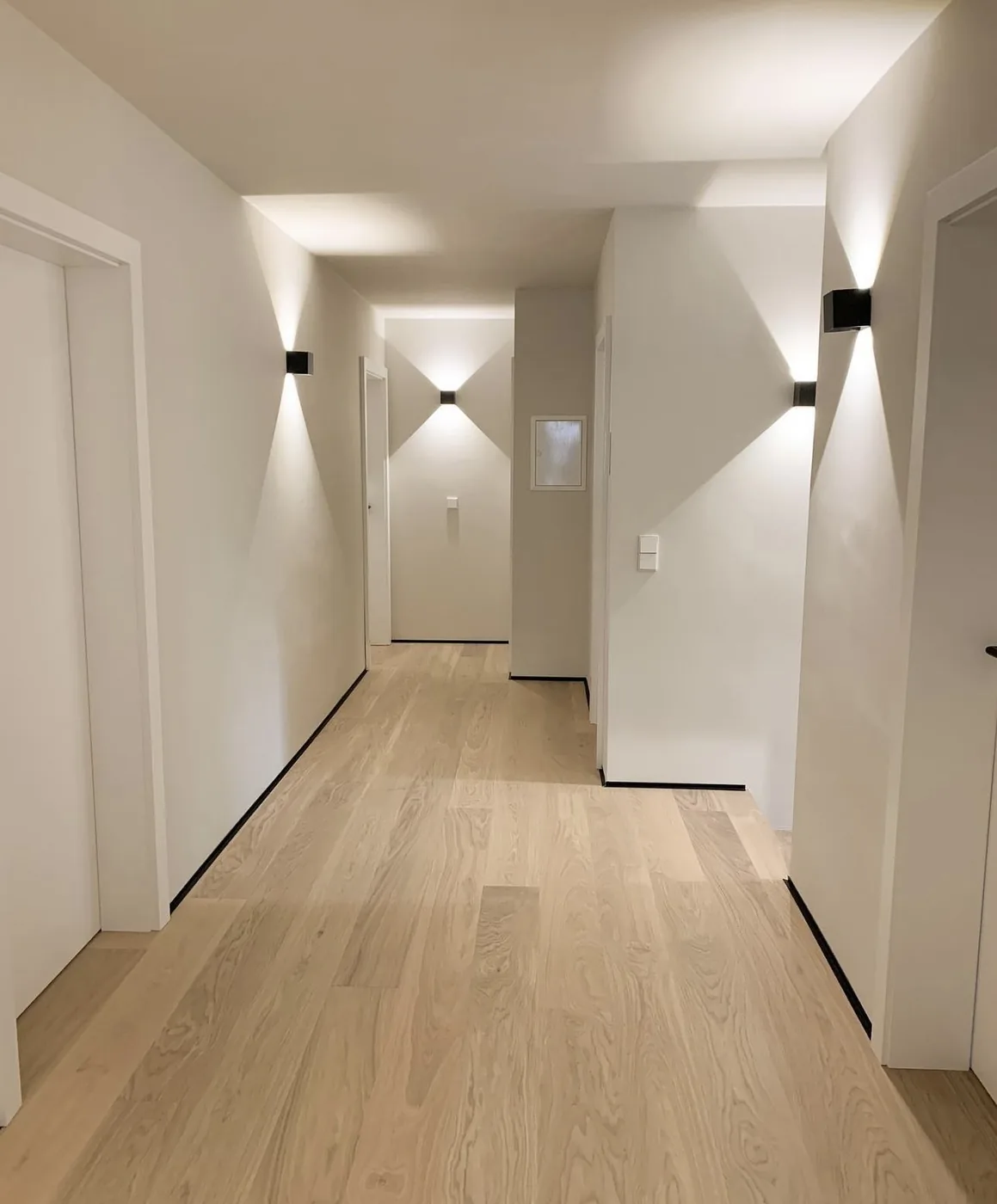 Light white oak plank floor in a long hallway with black baseboard reveals and wall lights, Toronto